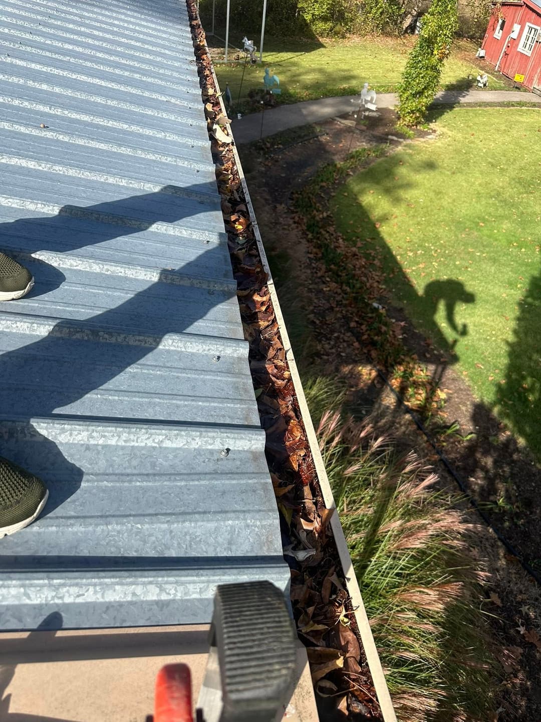 Person standing on a metal roof, clearing leaves from the gutter in a residential area.